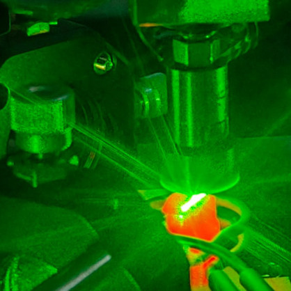 Das Bild zeigt grünes Laserlicht, das mit einem Mikroskopobjektiv auf eine rot-glühende Probe trifft.