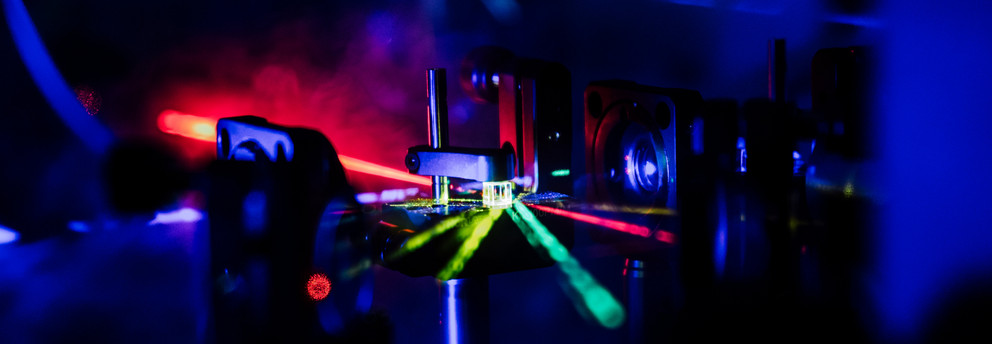 Das Bild zeigt mehrere, unterschiedlich farbige Laserstrahlen, die auf einen Strahlteilerwürfel treffen.