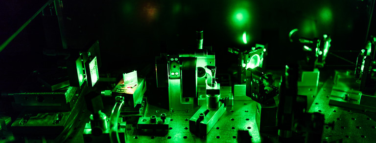 Das Bild zeigt grünes Laserlicht, das auf mehrere unterschiedliche optische Elemente trifft, in einem dunklen Labor.